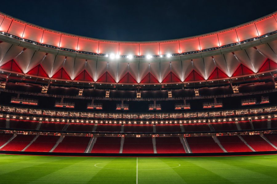 The empty bowl of a modern 70,000-seat football stadium at night, lit by floodlights