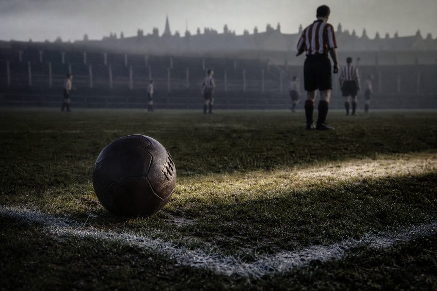 Old brown leather ball on floodlit grass beside a white line, a hazy stand behind
