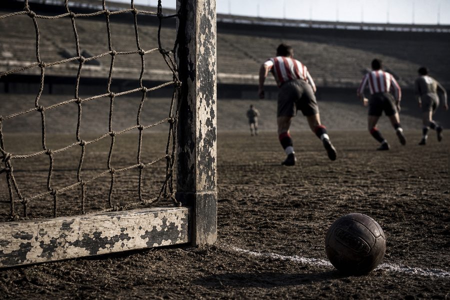 Peeling wooden goalpost and old net on a mud pitch, a leather ball on the line and players running away